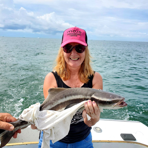 Bond woman in pink hat smiling holding a small cobia that is brown and looks like a catfish and about ten pounds. 