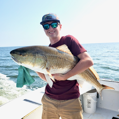 young adult in our cockpit holding a 35 pound redfish or red drum. sun shinning, scales look amber like a goldfish. Smiles. blue water.