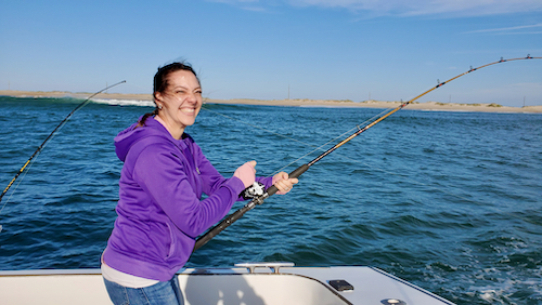smiling female angler in a purple coat. rod is bent over with a fish on it as the blue water and beach is seen in the foreground and background