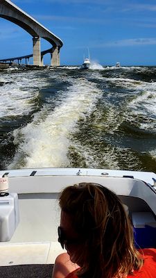 woman staring behind the boat with white caps and boat wake. Bigger boat approaching with a bridge to the left