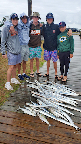 a group of five young men under twenty years of age, smiling, behind their catch of silver, ribbon fish on the dock