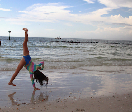 young girl with tye dye shirt in a bathing suit, doing a one handed cartwheel mid cartwheel on the beach. blue sky and ocean in background