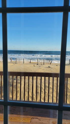 a sunny view out a window from an oceanfront home. blue skies, sandy beach, small waves on a blue ocean