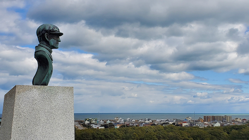 a Bronze Bust of Wilbur Wright of the Wright Brothers airplane which is on a cement pillar . His gaze overlooks the ocean and small cottages in the distance