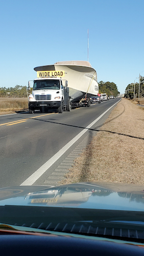 A truck carrying a biege boat with no cabin on it driving down the road. boat is very large. 6o ft. 