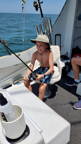 young boy sitting in the chair on the boat. he is smiling, wearing a sun hat and cranking in a fish! 